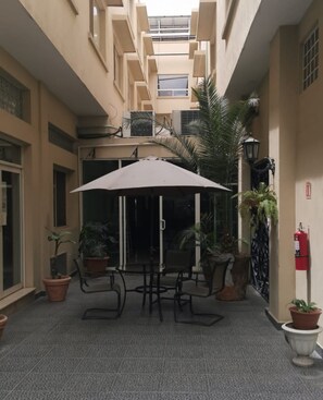 Une cour avec une table et des chaises placées sous un grand parasol, entourée de plantes en pot et agrémentée d’un extincteur fixé au mur.