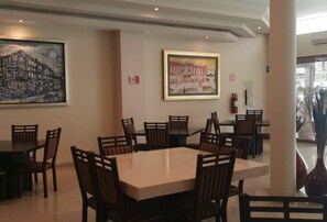 Un espace repas avec des tables et des chaises rectangulaires, deux tableaux accrochés aux murs et un extincteur.