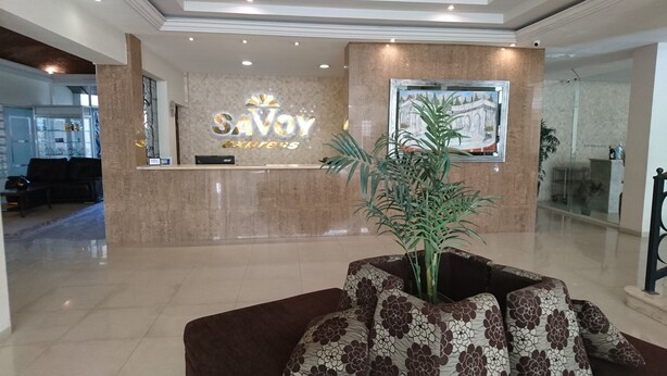 Zona de recepción con un cartel que dice 'Savoy', un mostrador de recepción, una planta en maceta y un cuadro en la pared.