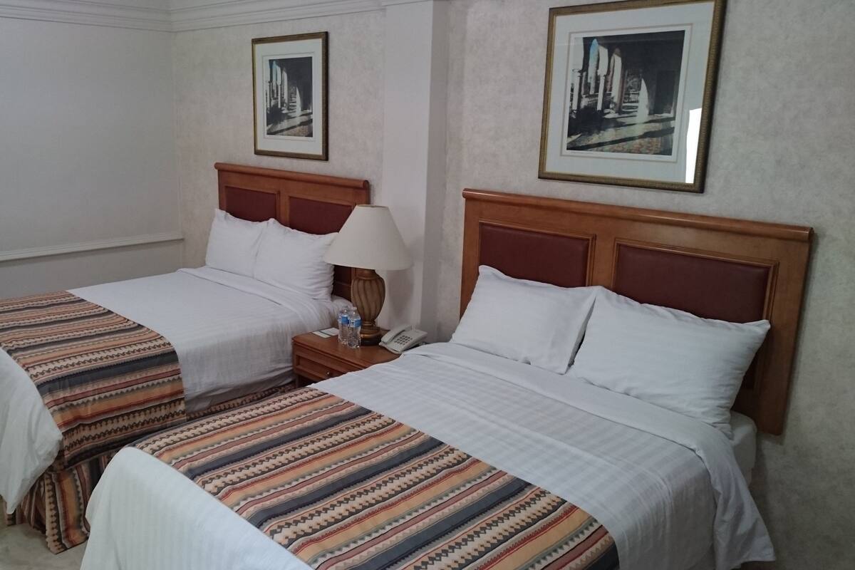 Ett hotellrum med två separata sängar, var och en med ett träfärgat sänggavel, vita sängkläder och en mönstrad pläd. Mellan sängarna står ett nattduksbord med en lampa och en vattenflaska.