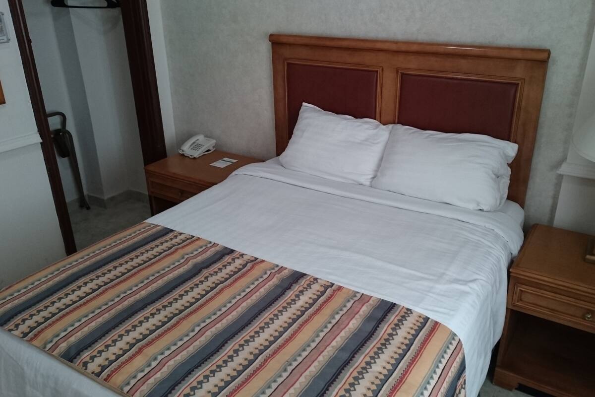 Ett hotellrum med en säng, ett nattduksbord med en lampa, en telefon och en väggmonterad lampa.