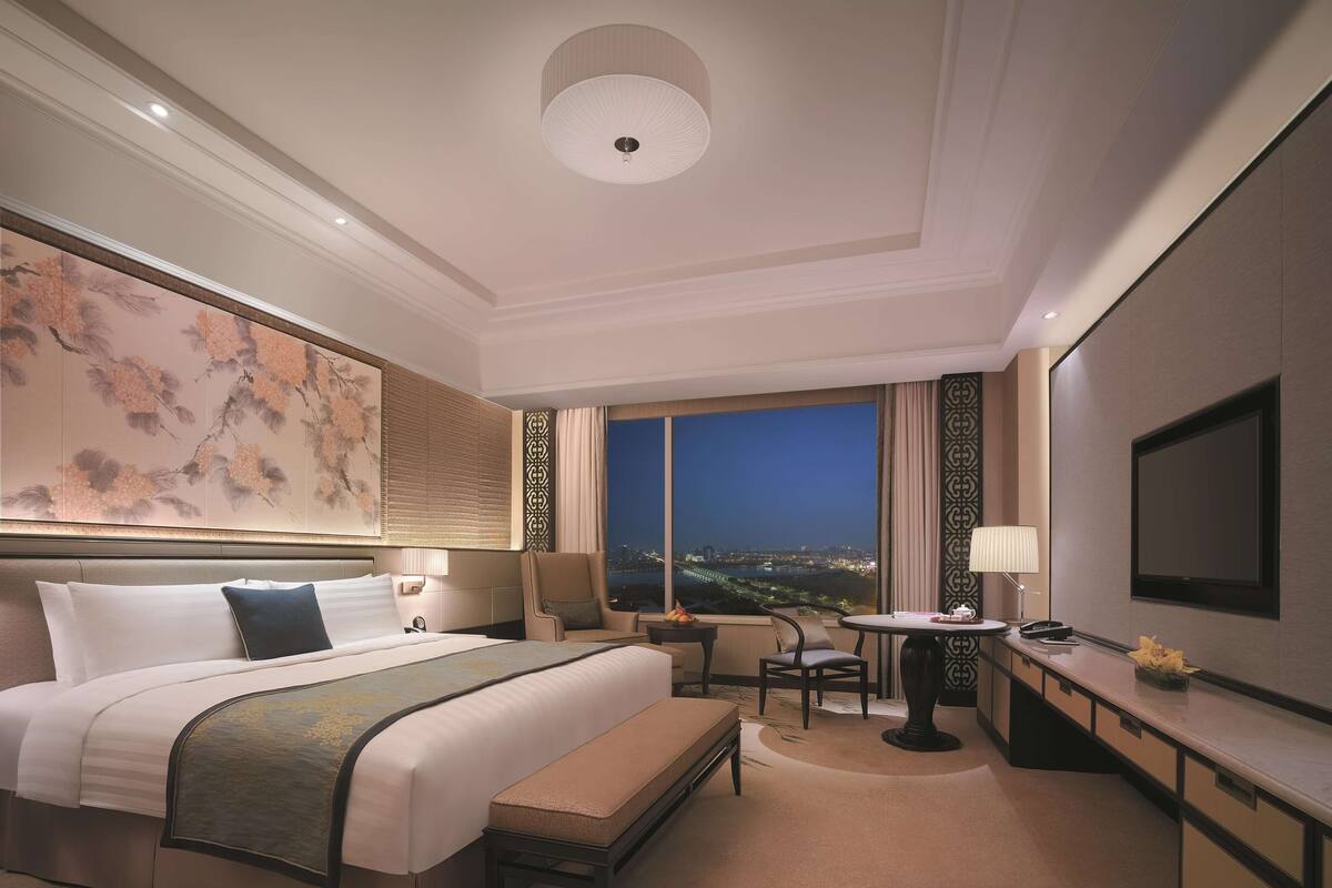 Une chambre d’hôtel avec un grand lit, un bureau, une chaise, une vue sur la ville la nuit et un téléviseur.