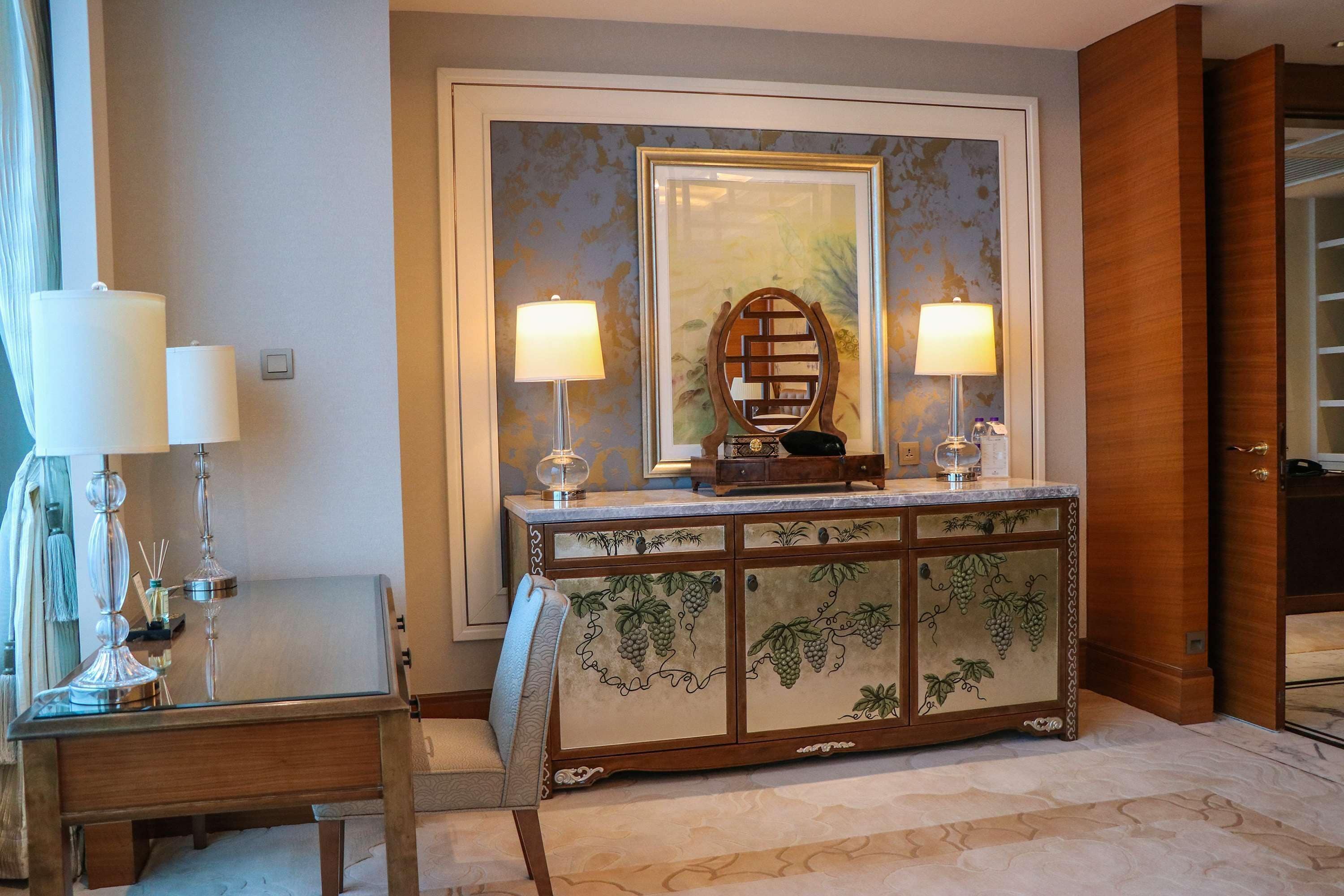Un hall de hotel con un mostrador de recepción de mármol, dos lámparas fijadas a la pared, un espejo grande enmarco y un mueble de madera con diseño de parra.