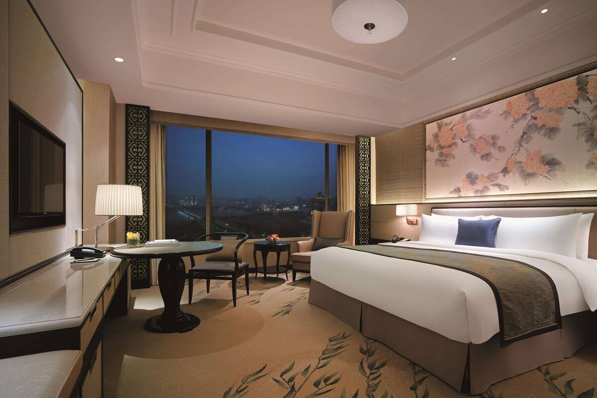 Une chambre d’hôtel avec un grand lit, un bureau, une chaise, une table et une vue sur la ville la nuit.