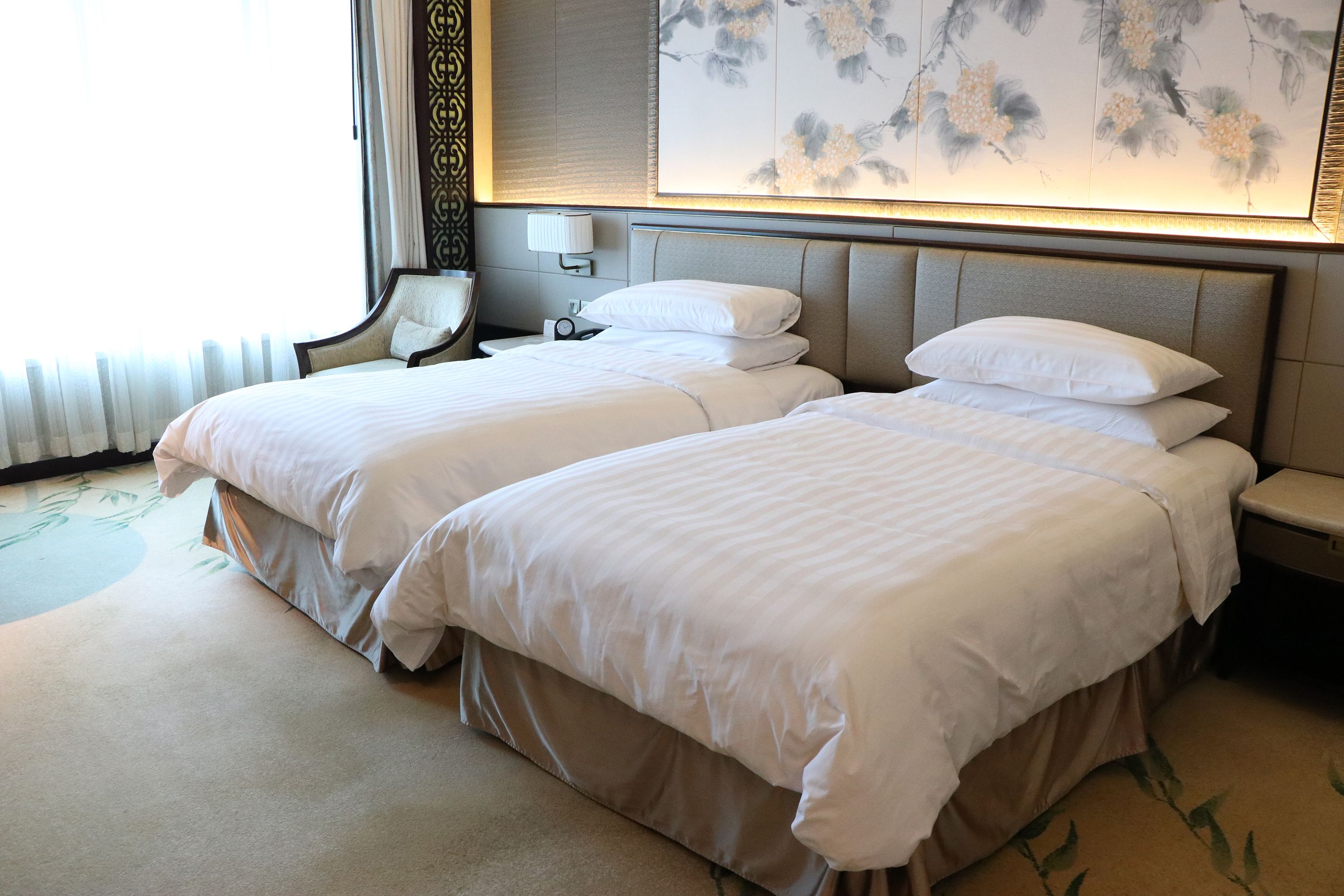 A hotel room with two beds, a bedside table, a lamp, and a floral artwork on the wall.