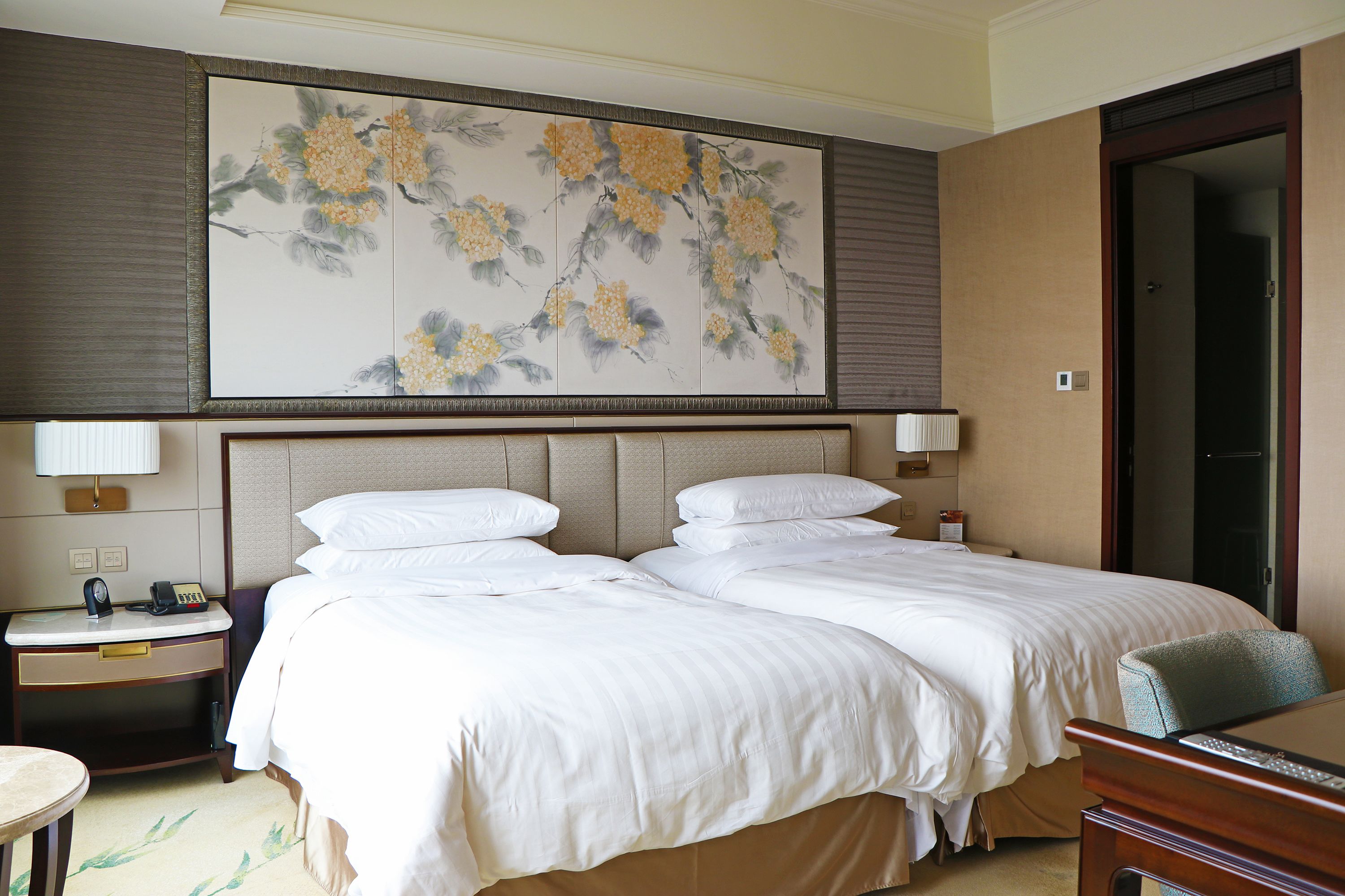 A hotel room with two beds, a large floral artwork above the beds, a nightstand with a lamp, and a desk with a chair.