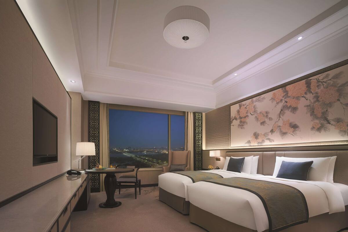 Une chambre d’hôtel avec un grand lit, un bureau avec une lampe, une chaise et une vue sur la ville la nuit.