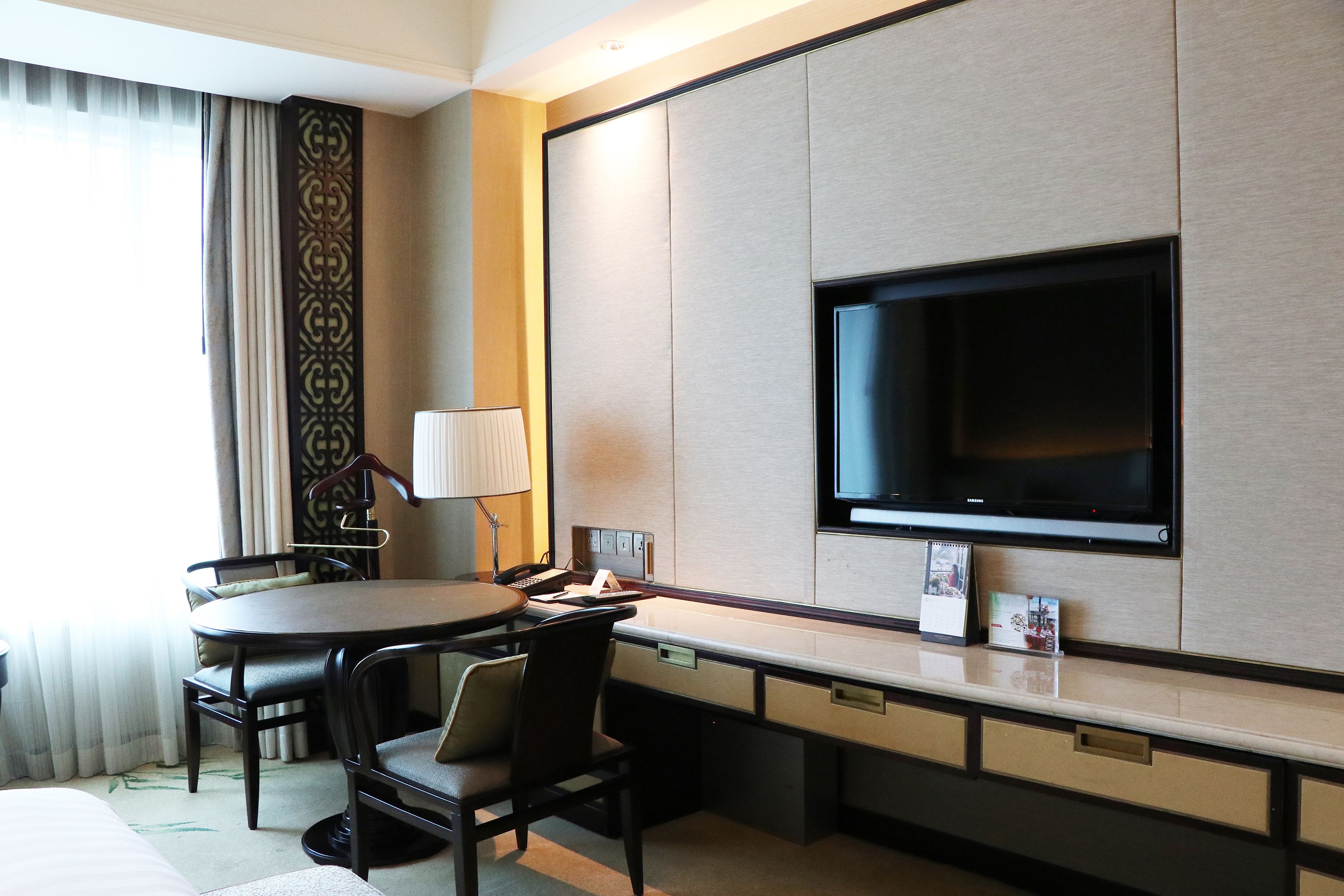 Habitación de hotel con una cama, un escritorio con silla, un televisor en la pared y una ventana con cortinas.
