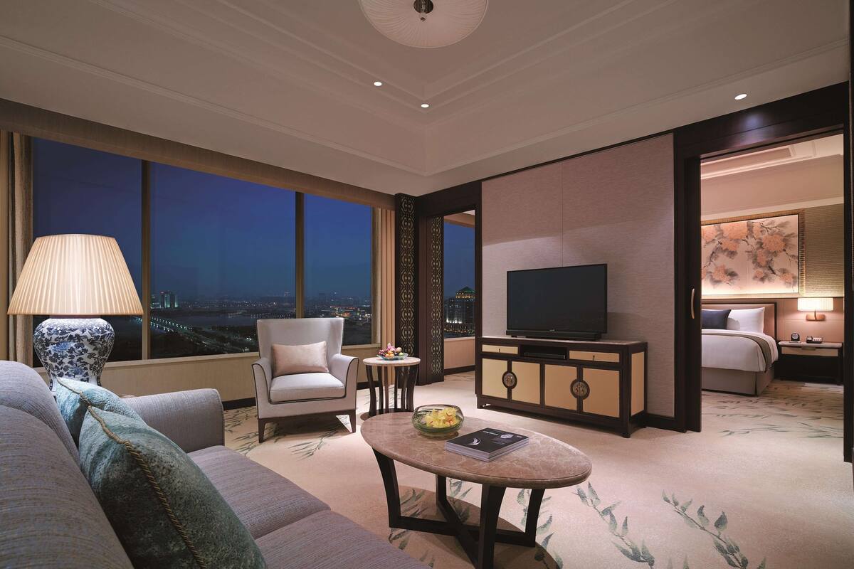 Une chambre d’hôtel moderne dotée d’une grande fenêtre, d’un téléviseur à écran plat, d’un canapé, d’une petite table ronde et d’un espace chambre séparé.