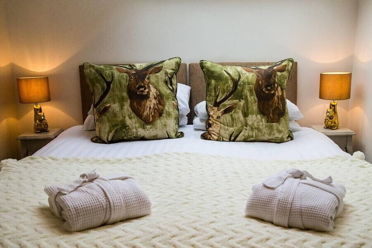 A neatly made bed with two decorative pillows featuring deer, a white textured bedspread, and two rolled towels on the bed.