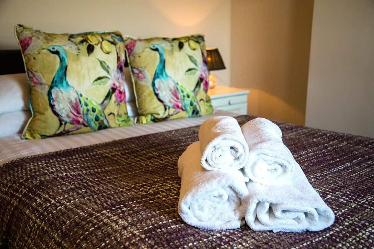 A neatly made bed with two pillows featuring a bird and floral design, and two rolled towels on the bed.