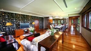 Uma sala de estar ampla, com piso de madeira, um sofá, uma mesa de centro e uma televisão. Há também uma área de jantar com uma mesa comprida e cadeiras.