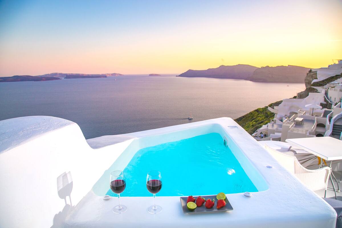 A poolside view with a sunset over the sea, white buildings, and a table with drinks and fruit.
