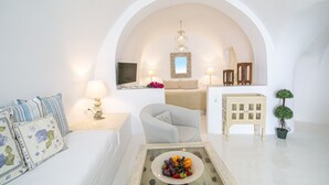 A white-themed living room with a sofa, armchair, and a coffee table with fruit. There is a TV, lamps, and a mirror on the wall. - Onar Villas Oia - Onar Hotels Collection (Santorini)