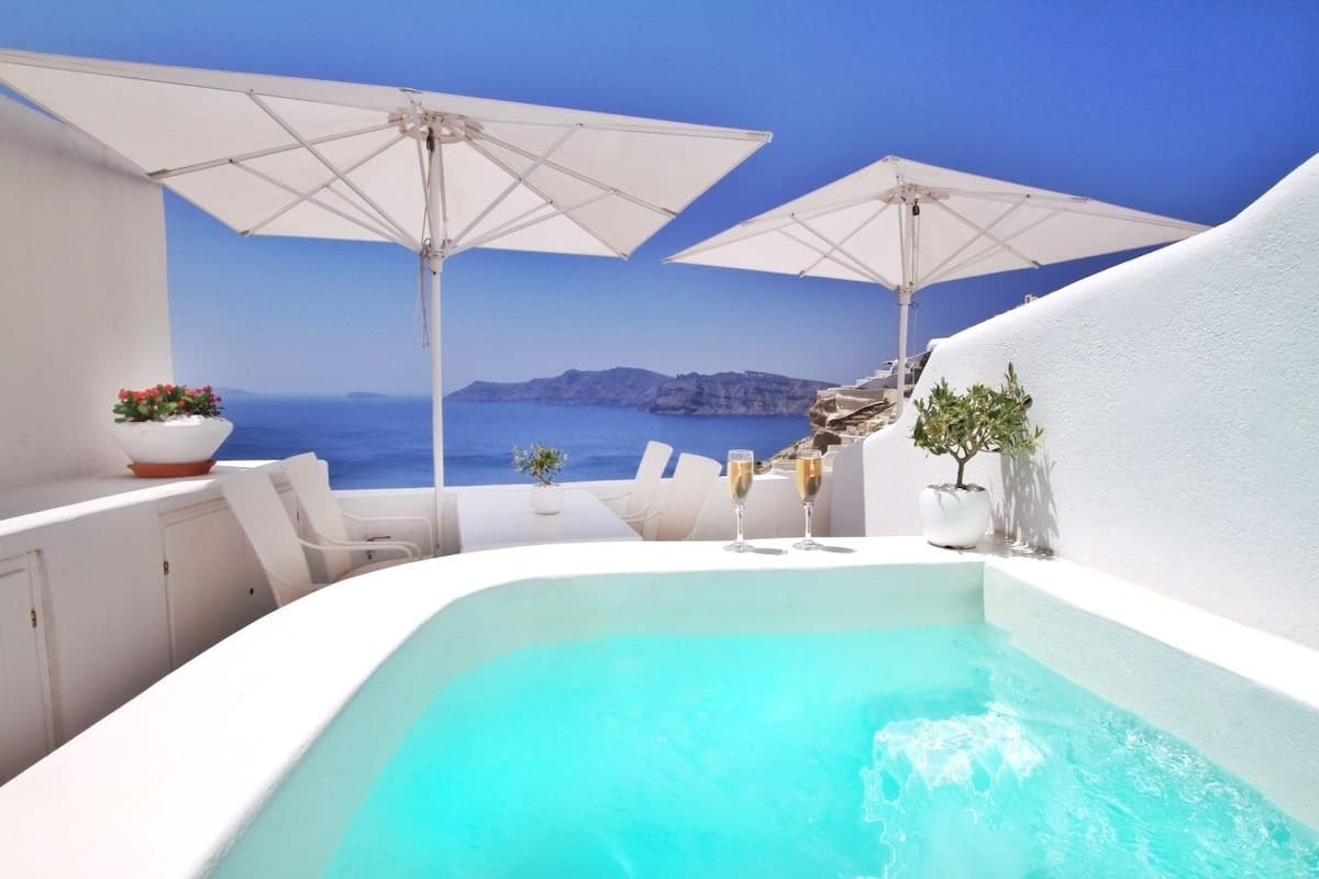 A white infinity pool with two white umbrellas, a white table with chairs, and a view of the sea.