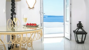 A room with a glass table, two glasses of white wine, a plate with strawberries, a black lantern, and a view of the sea through an open door. - Onar Villas Oia - Onar Hotels Collection (Santorini)