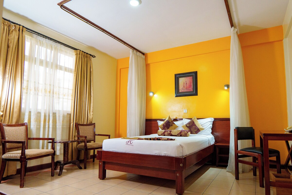 A hotel room with a bed, two chairs, a desk, and a painting on the wall.