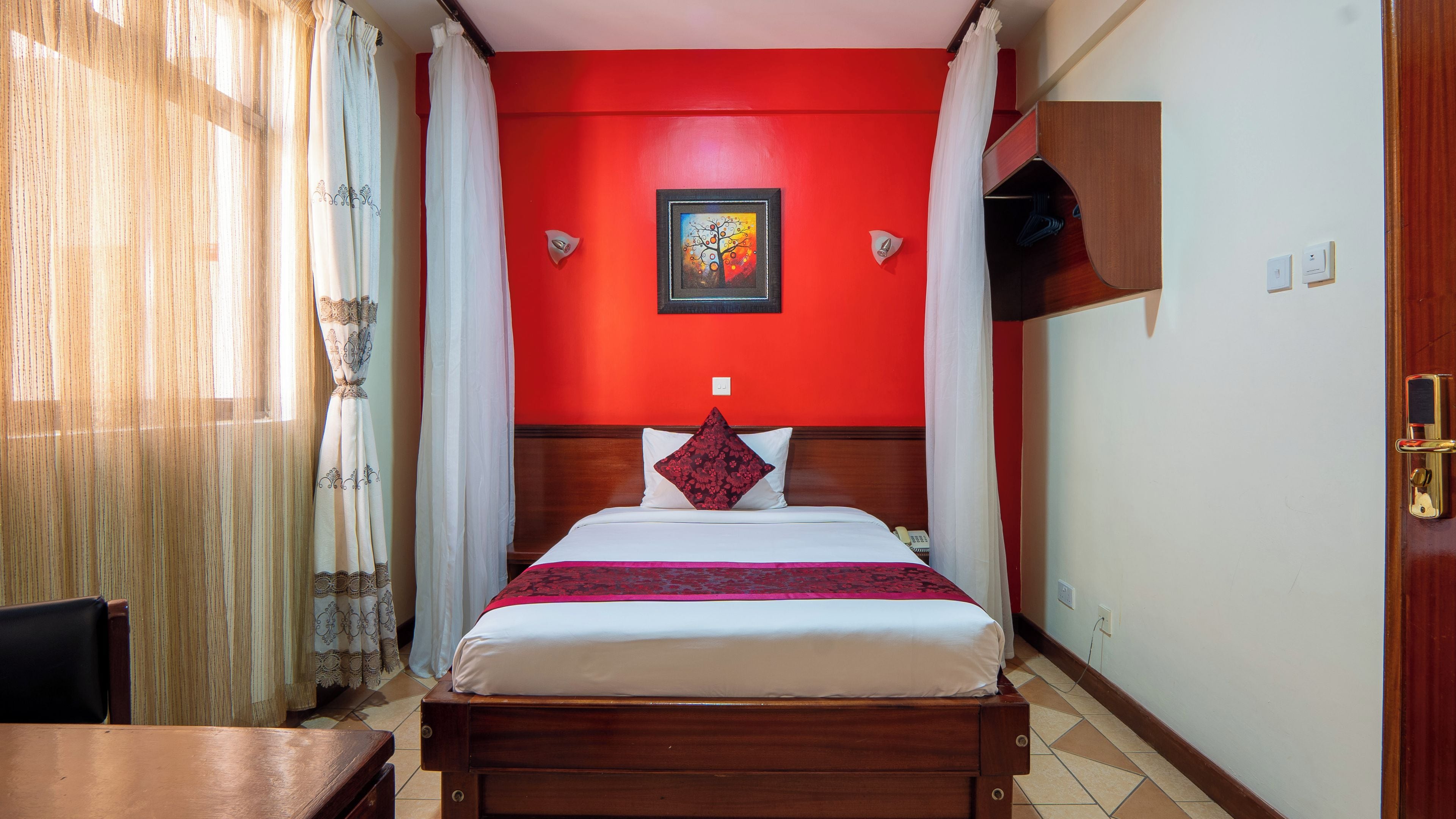 Une chambre d’hôtel avec un lit, une tête de lit en bois, un mur rouge, un tableau et une fenêtre avec des rideaux.
