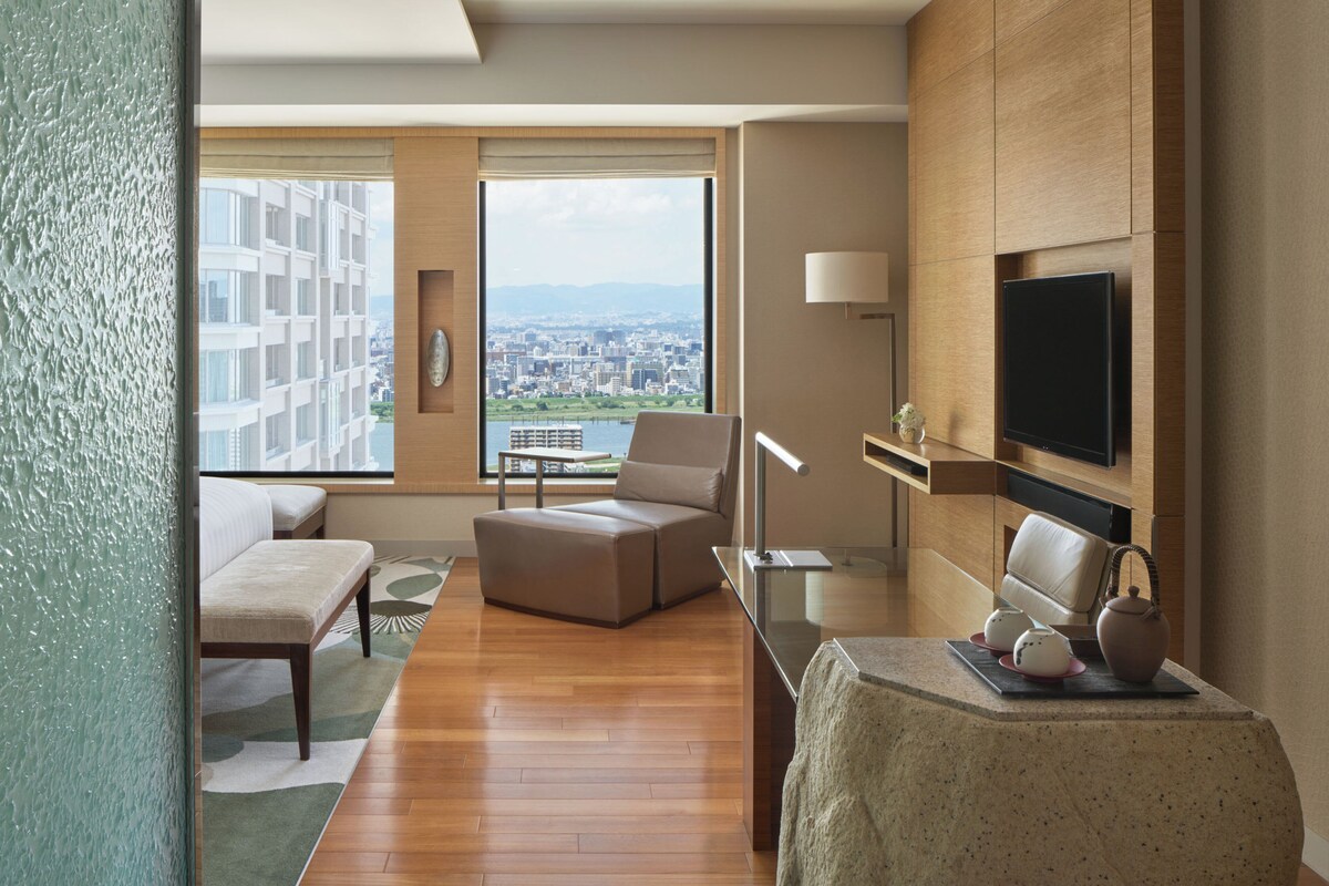A modern hotel room with a large window offering a city view, a flat-screen TV, a comfortable seating area, and a small table with decorative items.