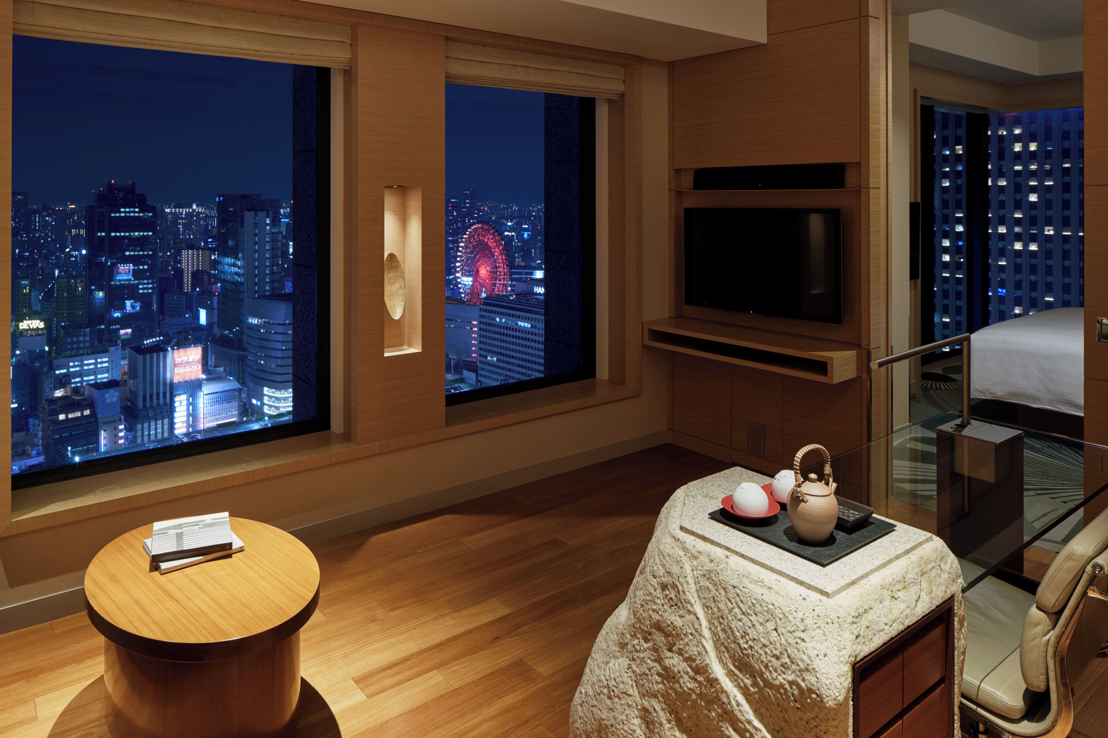 A hotel room with a large window offering a city view, a flat-screen TV, a small round table, and a bed with a glass-enclosed balcony.