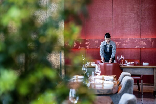 A restaurant interior with a chef preparing food at a counter, a dining table set with plates and glasses, and a red wall with a subtle pattern. - InterContinental Osaka by IHG (Osaka)