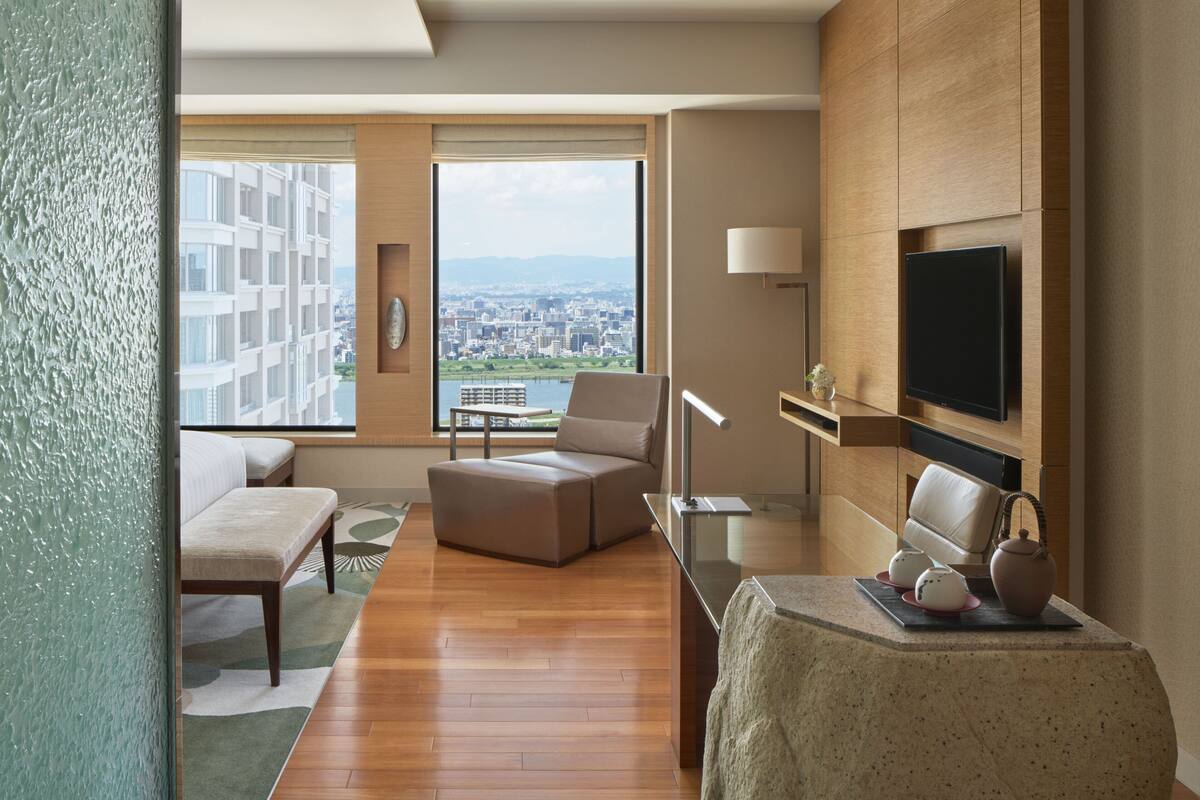 Habitació d'hotel moderna amb una finestra gran que ofereix vista a la ciutat, una televisió de pantalla plana, zona d'estar còmoda i una taula petita amb objectes decoratius.