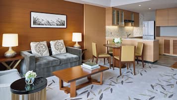 A modern hotel room with a grey sofa, two lamps, a wooden coffee table, and a dining area with chairs and a small island.