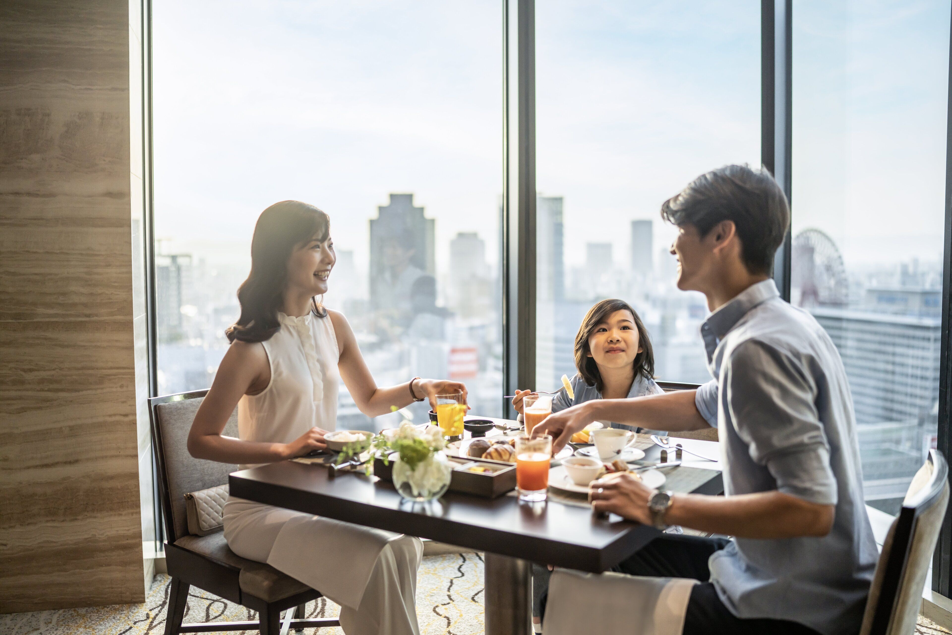A group of people dining at a table with a city view, featuring a modern interior with large windows and a cityscape in the background.