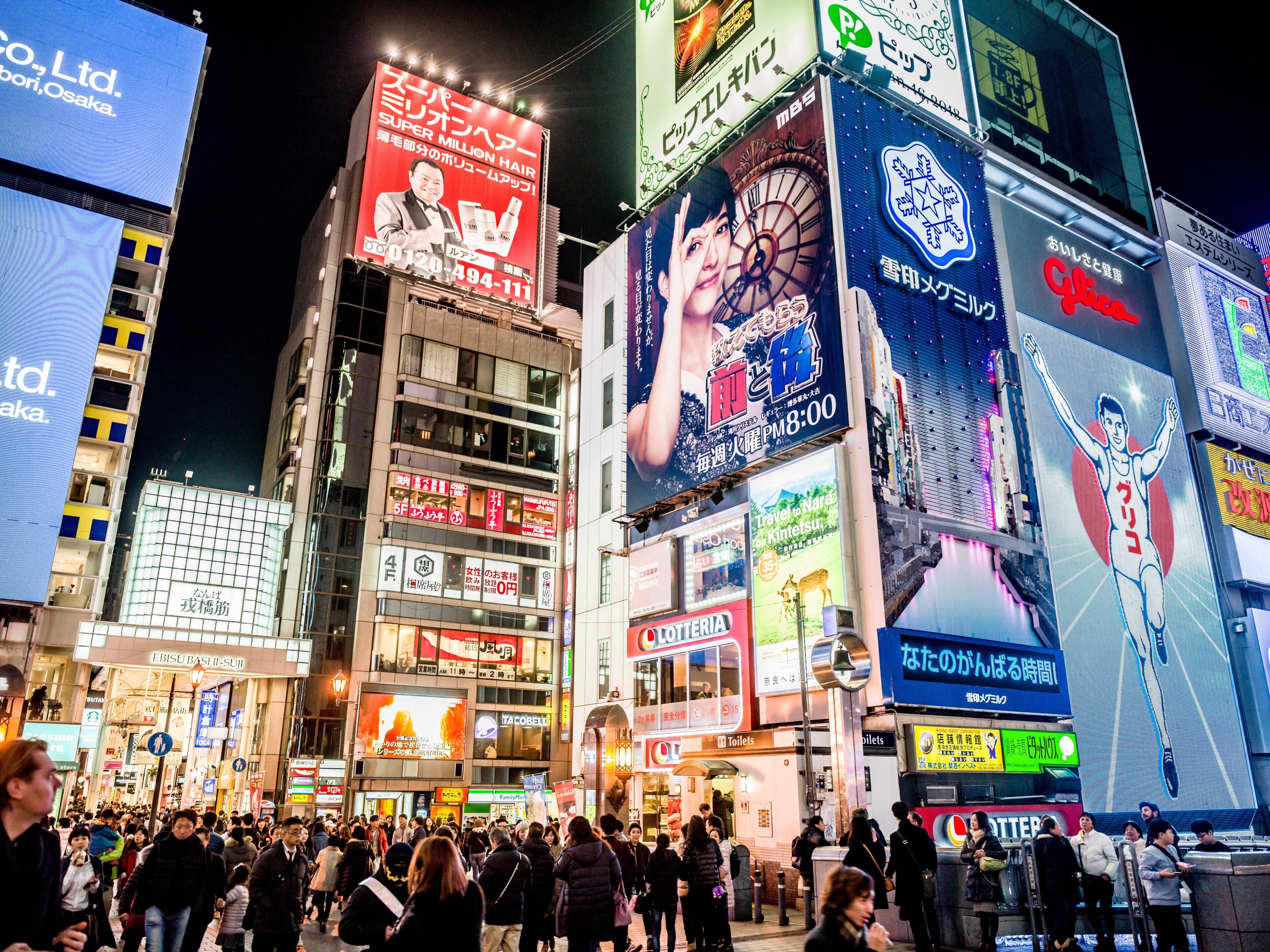 A busy urban street at night with illuminated billboards and a crowd of people.