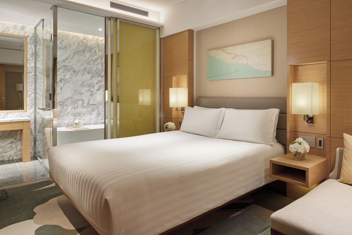 A hotel room with a large bed, a glass shower enclosure, a wooden nightstand with a lamp, and a wall-mounted map.