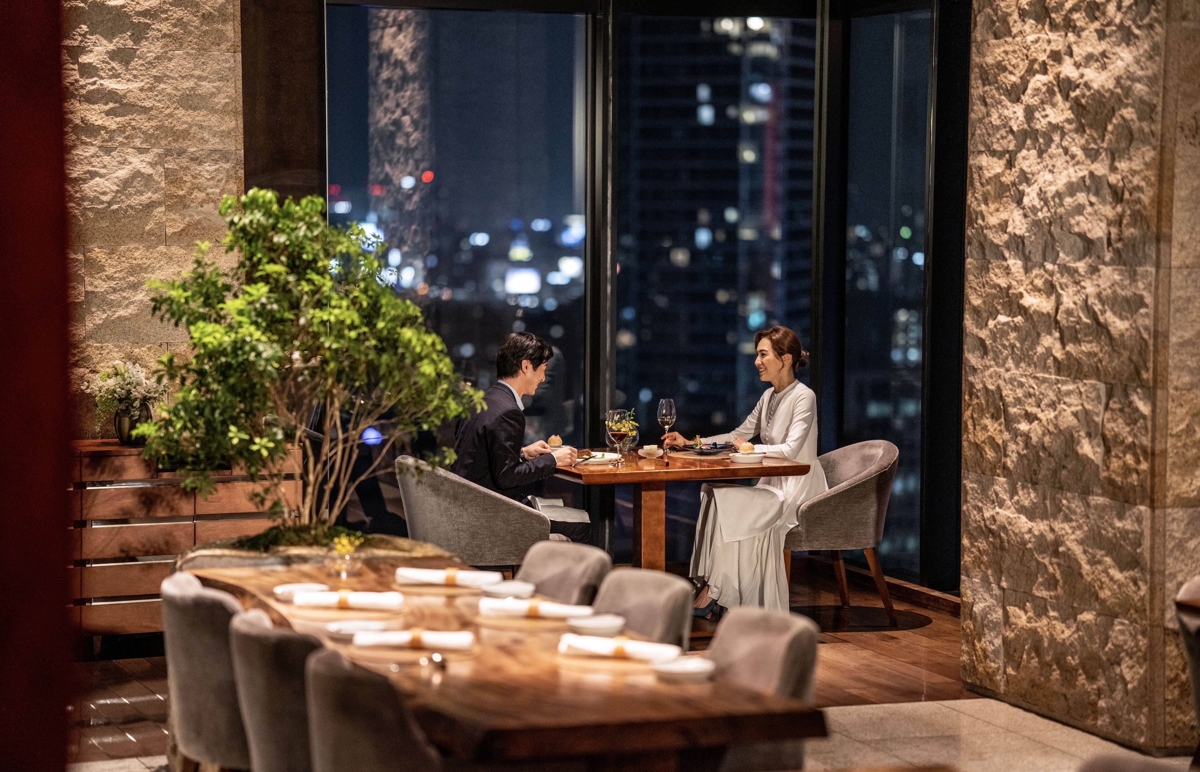 A dining area with a table set for two, a city view through large windows, and a stone wall.