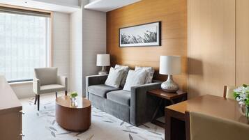 A modern hotel room with a grey sofa, two armchairs, a wooden coffee table, and a wooden accent wall with a framed picture.