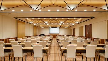 A conference room with rows of tables and chairs, a large screen at the front, and a geometric ceiling design.
