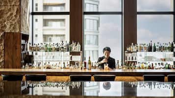 A bar with a bartender, shelves of liquor bottles, and a large window with a city view.
