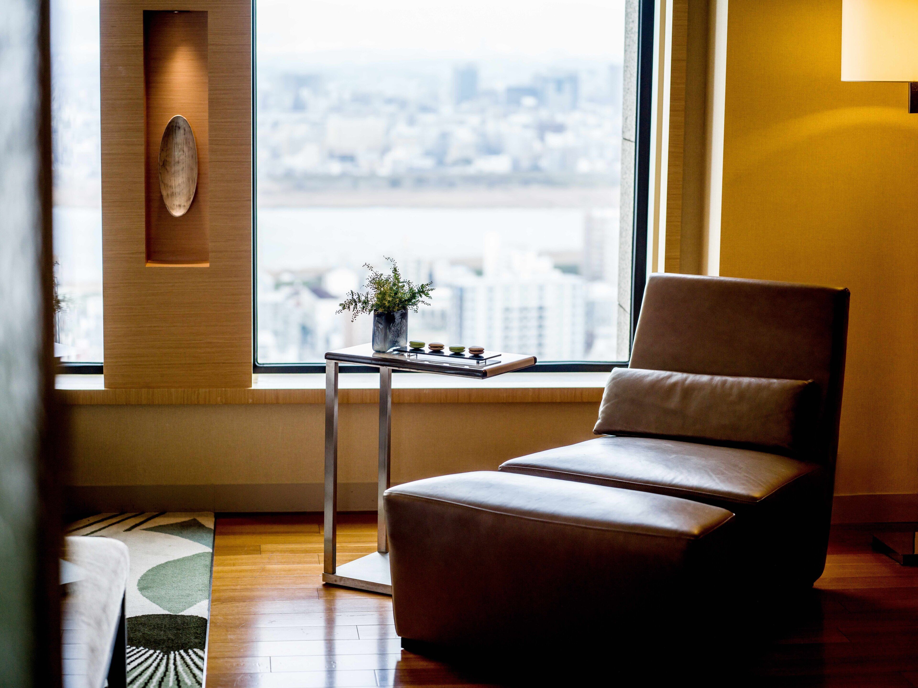A modern room with a leather chair, a small table with a potted plant, and a view of a cityscape through a large window.