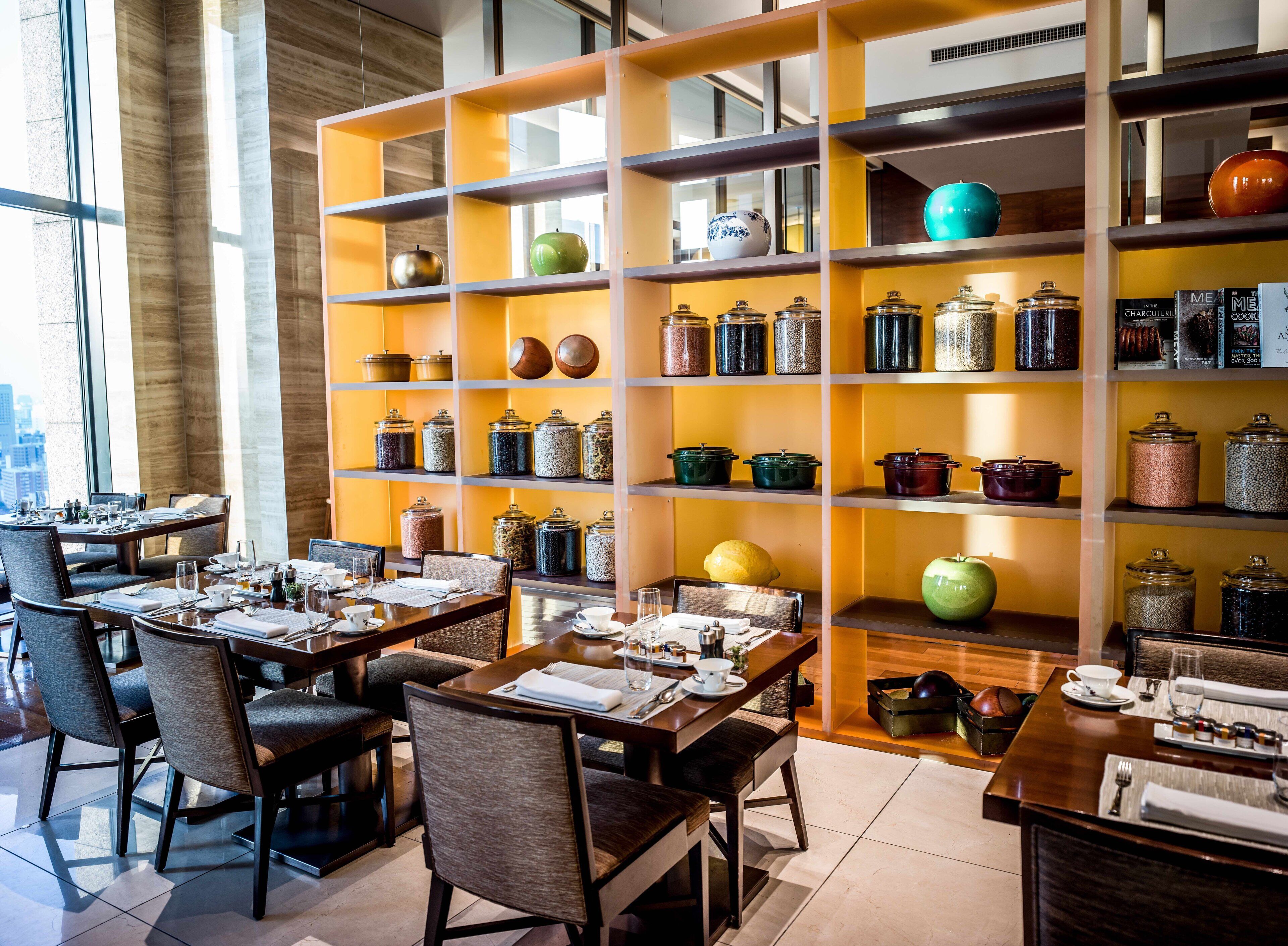 A restaurant with multiple tables and chairs, a large shelf with jars and kitchenware, and a view of the city through large windows.