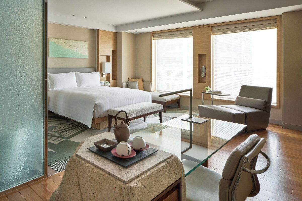 A hotel room with a large bed, a seating area with a sofa and armchair, a small table with a vase and teapot, and a window with a view.