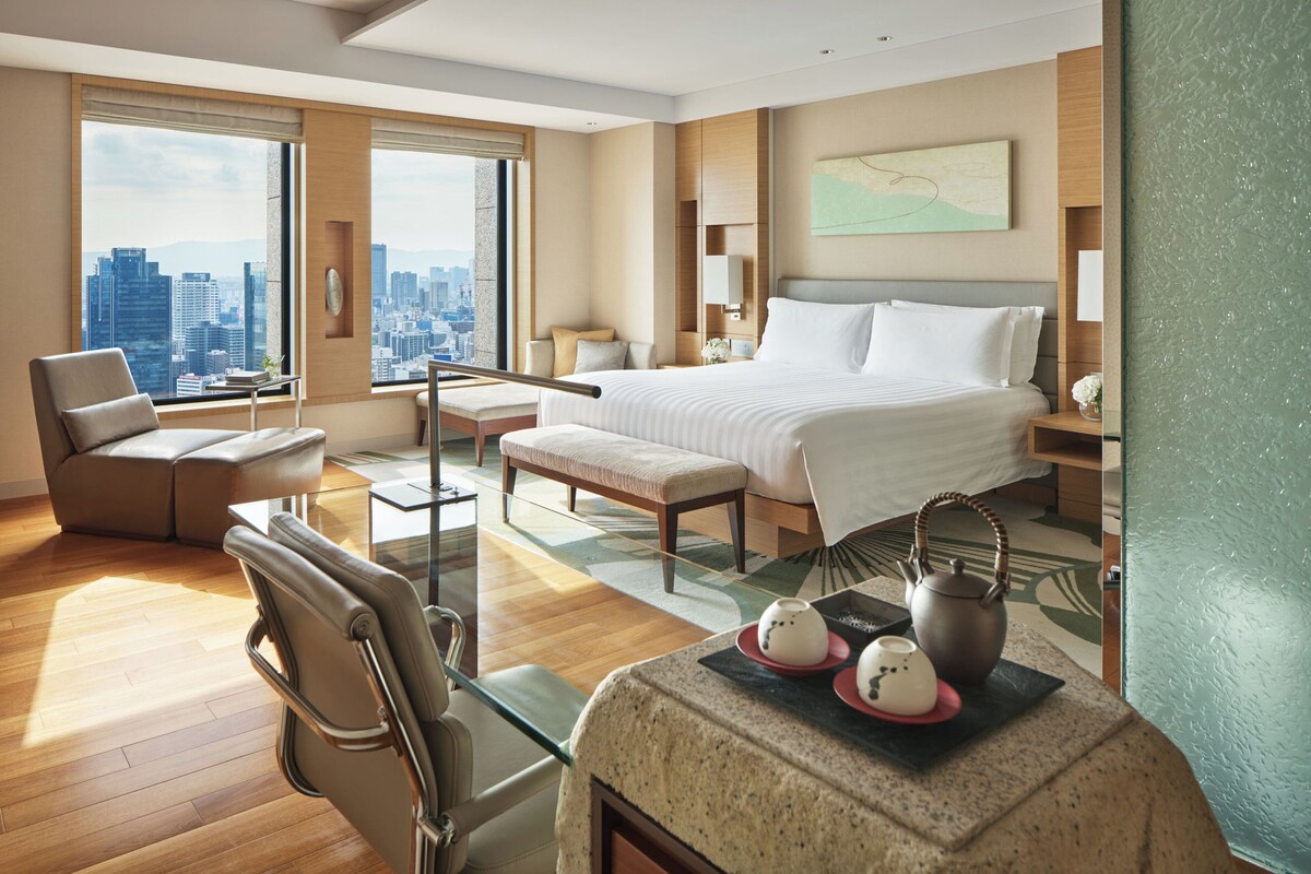 A modern hotel room with a large bed, a sitting area with a chair and a bench, a small table with a teapot and cups, and a city view through large windows.
