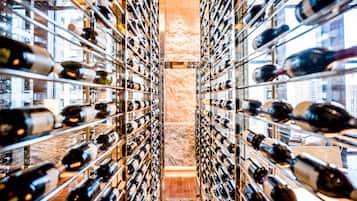 A wine cellar with rows of wine bottles on shelves, a stone wall, and a view of buildings through a glass door.