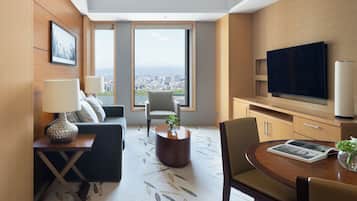 A modern hotel room with a city view, a flat-screen TV, a dining area, and a seating area with a sofa and armchairs.