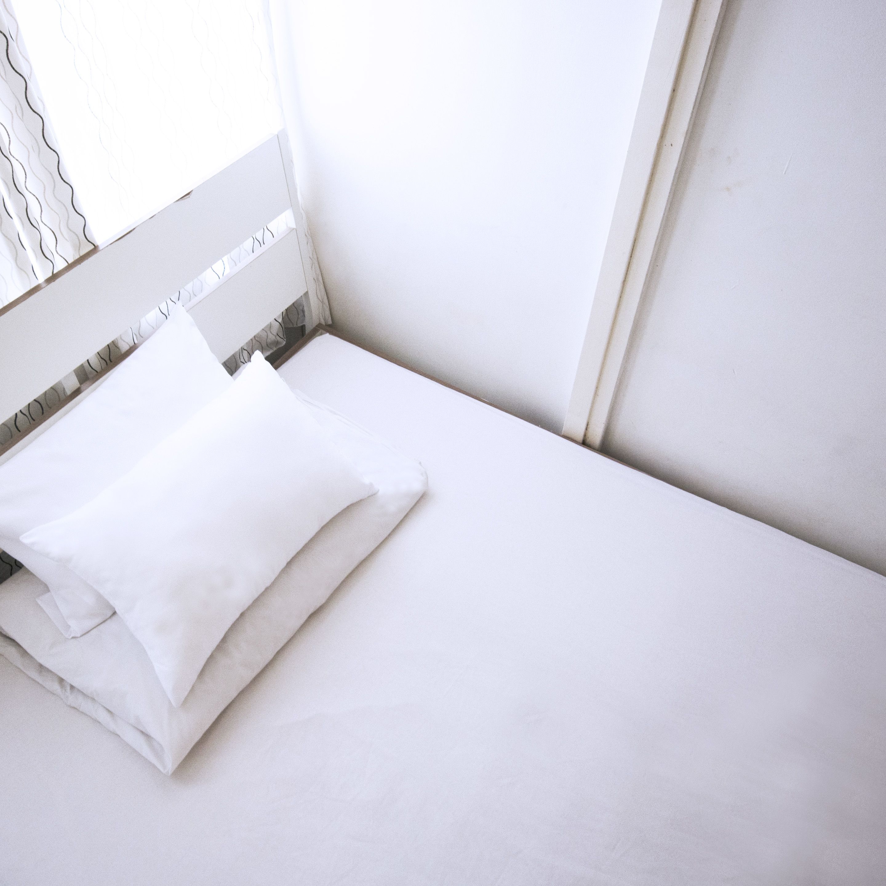 A neatly made bed with white bedding in a room with white walls and a window with a white curtain.