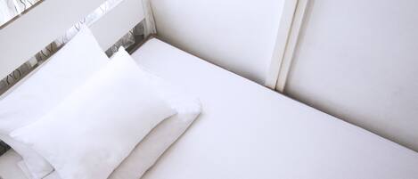 A neatly made bed with white bedding in a room with white walls and a window with a white curtain.