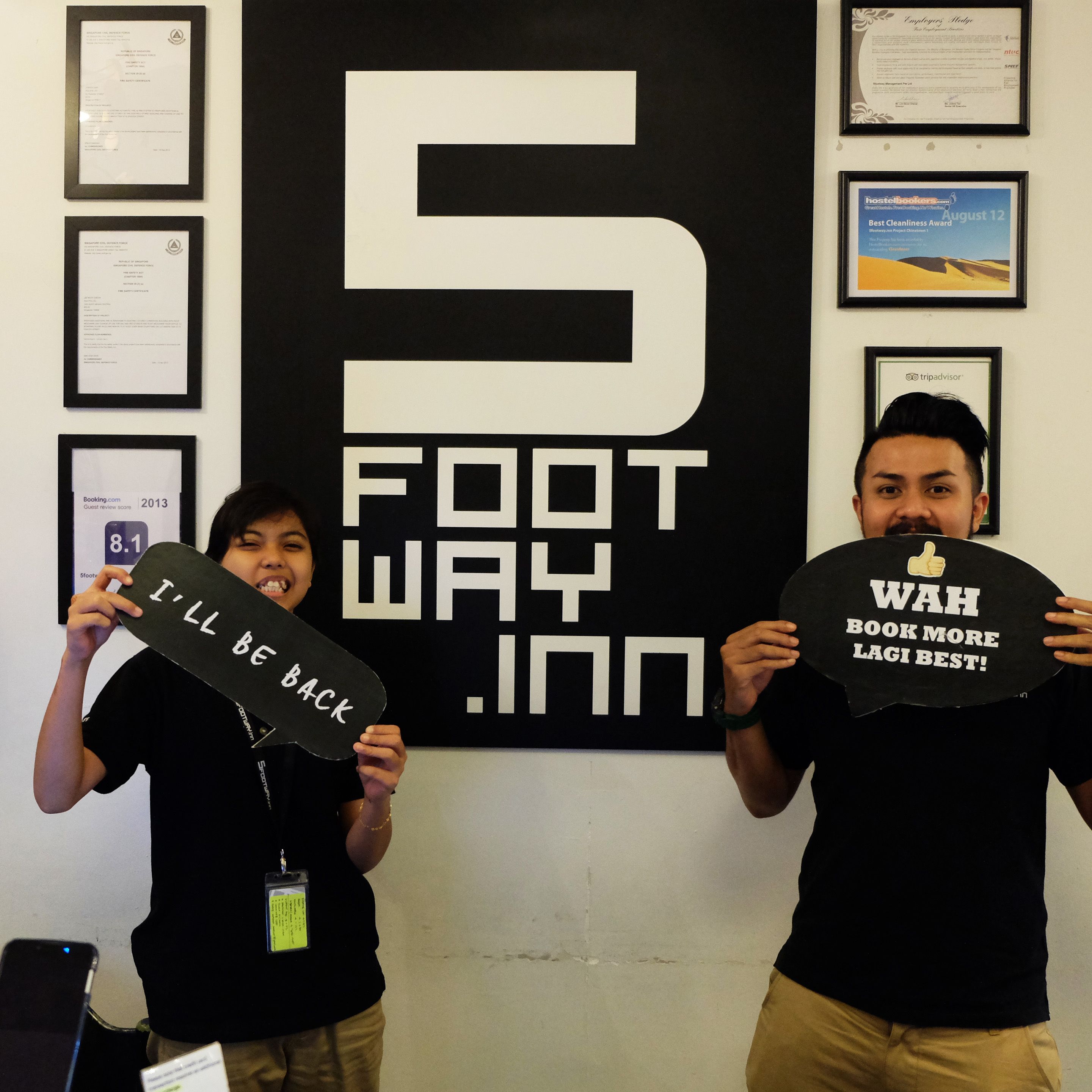 Two people holding signs in front of a wall with framed certificates and a large '5 FOOT WAY INN' sign.