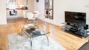Ein modernes Wohnzimmer mit einem Glastisch, weißen Sesseln, einem Flachbildfernseher und einer Küche im Hintergrund.