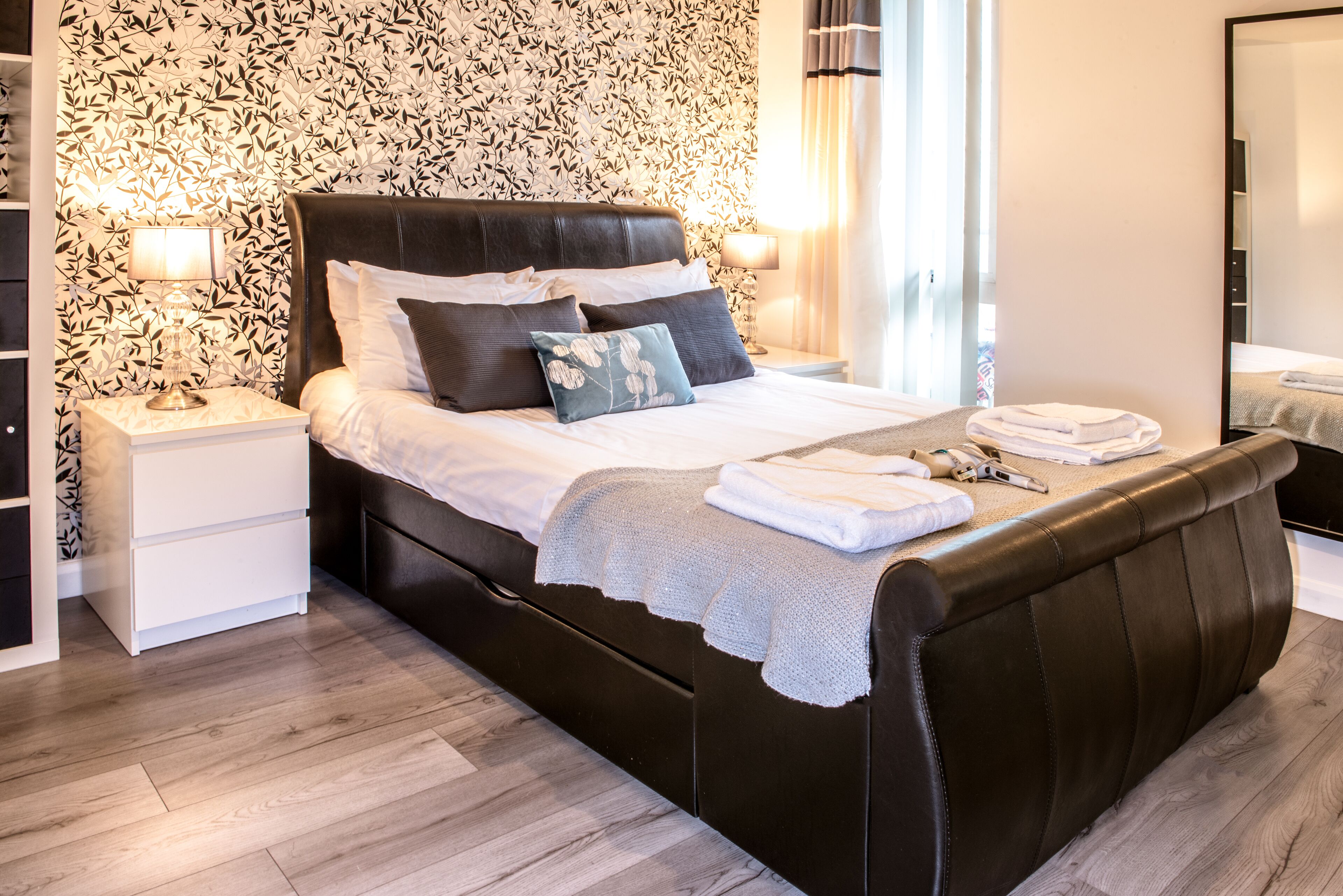 Un dormitorio moderno con una cama de cuero oscura, ropa de cama blanca y un papel tapiz estampado.