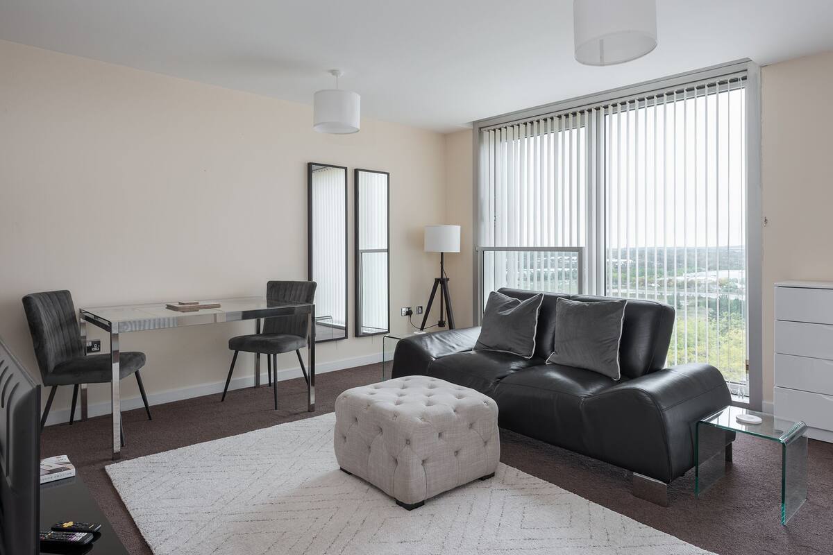 Un salon moderne avec un canapé en cuir noir, un pouf blanc, une table basse en verre et un coin repas comprenant un bureau blanc et deux chaises.