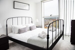 Een netjes opgemaakt bed met witte lakens en een grijze plaid, een nachtkastje met een lamp, en een raam met jaloezieën.