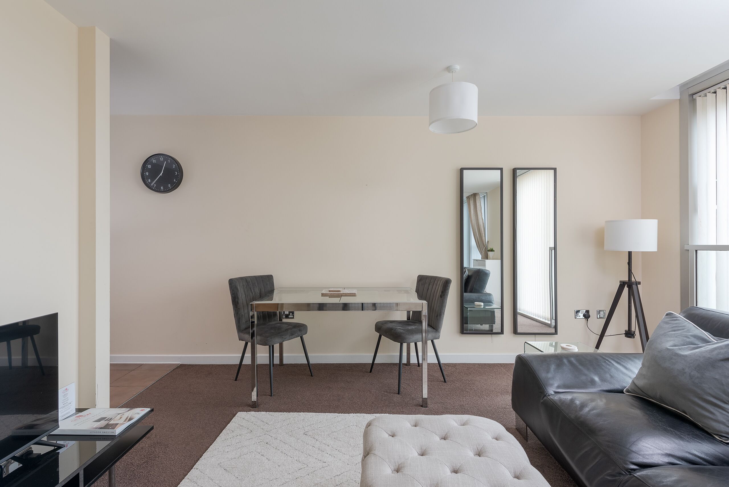 Een moderne woonkamer met een bank, een voetenbankje, een eettafel met stoelen, een televisie, een klok en twee spiegels.