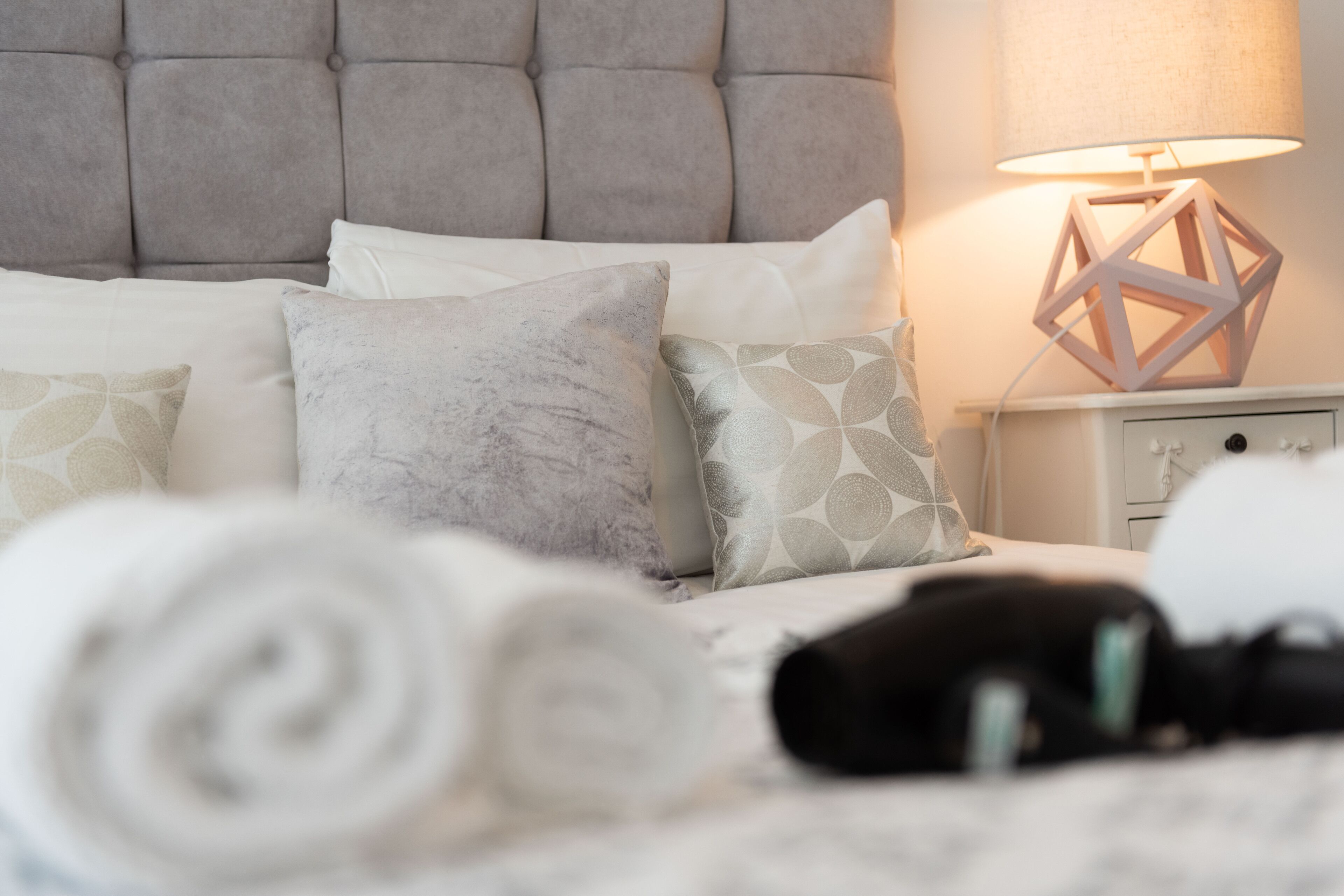 Una cama bien tendida con almohadas y una toalla blanca enrollada. Un objeto negro, posiblemente un secador de pelo, está sobre la cama. Una lámpara de noche con un diseño geométrico está sobre la mesita de noche.
