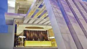 Et moderne højhushotel med en markant glasfacade, et receptionsområde med en gul disk og en tydelig trappe, der fører til de øvre etager.